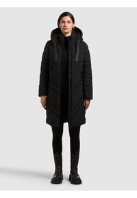 Sort pufferjakke med hætte, der har et chevron-design og frontlynlås. Båret med sorte leggings og kraftige sorte støvler.