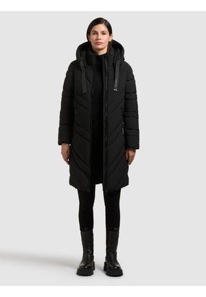 Sort pufferjakke med hætte, der har et chevron-design og frontlynlås. Båret med sorte leggings og kraftige sorte støvler.