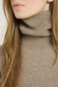 Maglione color beige con collo a costine, lavorazione a maglia testurizzata e tessuto morbido, indossato con capelli lunghi e ondulati che incorniciano il viso.