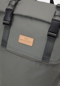 Grauer Canvas-Rucksack mit einer Roll-Top-Design, schwarzen Beschlägen und einem hellbraunen Lederetikett mit Text. Verstellbare Schultergurte für Bequemlichkeit.