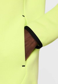 Prenda de alta visibilidad en color amarillo neón con un puño negro, que cuenta con un bolsillo y sutiles acentos de costura. Textura suave y diseño ajustado.