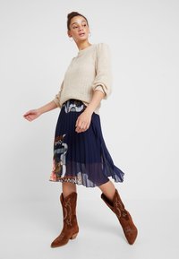 Maglione beige a maglia con maniche a sbuffo, gonna plissettata blu navy con motivi floreali e stivali da cowboy in suede marrone con cuciture dettagliate.