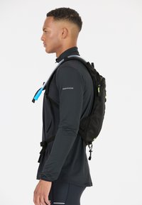 Black hydration backpack with mesh panels, adjustable straps, and a blue hydration tube. Model wears a black long-sleeve top.