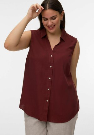 Mujer sonriente ajustándose el cabello, vestida con una camisa sin mangas burgundy abotonada y pantalones beige claro, con un fondo sencillo.