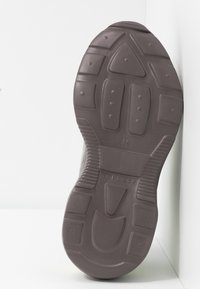 Semelle d'une chaussure marron foncé avec un motif de bande de roulement texturé et un marquage "TOPSHOP", montrée sur une surface blanche et dans un coin de mur.