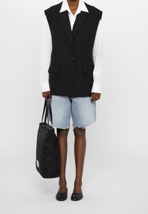 Blazer noir sans manches avec deux poches avant porté sur une chemise blanche, associé à un short en denim bleu clair et des ballerines noires, tenant un sac fourre-tout noir.