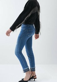 Niebieskie dżinsy z przetarciami i frędzlami na dole, zestawione z czarną kurtką z frędzlami i czarnymi butami na obcasie typu slingback.
