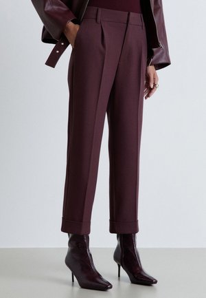 Donna che indossa pantaloni stretti color bordeaux scuro con risvolti, abbinati a stivaletti aderenti coordinati e una manica di giacca in pelle visibile.