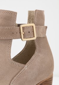 Bottine en daim marron clair dotée d'un design découpé, d'une boucle dorée et de coutures visibles. Le talon est en bois avec une finition texturée.