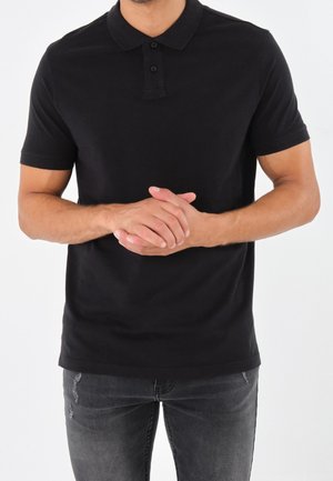 Mann trägt ein schwarzes kurzärmeliges Poloshirt und dunkelgraue Jeans, steht mit verschränkten Händen vorne vor einem schlichten weißen Hintergrund.