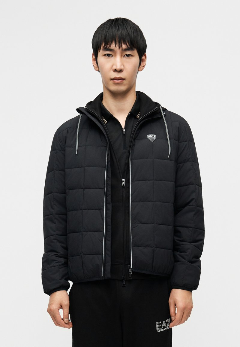 Μαύρο μπουφάν με καπιτονέ σχέδιο, που διαθέτει φερμουάρ στο μπροστινό μέρος και κουκούλα με ρυθμιζόμενο κορδόνι. Περιλαμβάνει λογότυπο στο στήθος και ελαστικά μανίκια.