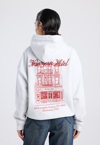 Persona con cabello corto y oscuro, vistiendo una sudadera gris claro con un dibujo rojo de un edificio y el texto "Hotel Nueva York, 245 Lexington Avenue, Manhattan, EE. UU."