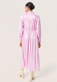 Abito rosa a maniche lunghe con schiena arricciata, colletto bottonato e gonna fluida. La modella indossa stivali beige fino al ginocchio.