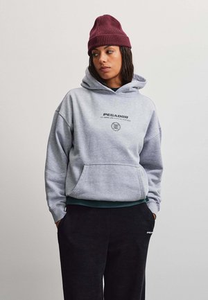 Giovane donna con indosso un berretto bordeaux, una felpa grigia con cappuccio con il testo "PEGADOR" e pantaloni neri con le mani nelle tasche, che guarda a sinistra.