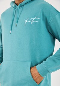 Persona con sudadera color verde azulado y texto blanco "Tu turno" en el pecho, con las manos en el bolsillo delantero y los cordones del mismo color visibles.