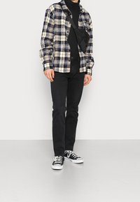 Rutig flanellskjorta i marinblått och krämvitt, svart polotröja, svarta jeans och svarta sneakers med vita sulor och snören. Detalj av axelremsväska.