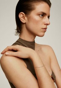 Pulsera de plata en una muñeca, contra un hombro desnudo y una tela de color verde oliva. Textura suave, diseño delicado, brillo sutil.