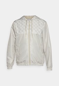 Chaqueta cortavientos de color beige claro con una sección superior estampada, cierre completo, capucha ajustable y puños y dobladillo elásticos. Textura suave en toda la prenda.