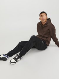 Sudadera marrón con cordones y logo, combinada con pantalones de chándal negros y zapatillas blancas con detalles en azul marino, sentada contra un fondo blanco.