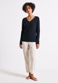 Marineblauwe V-hals trui met lange mouwen, gecombineerd met lichtbeige geruite broek en tan platte sandalen. Gladde stoftexturen, ontspannen pasvorm.