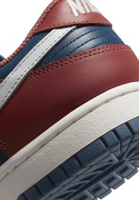 Gros plan du talon d'une basket en cuir bleu et rouge avec des coutures blanches, une semelle intermédiaire blanche, une semelle extérieure bleue texturée et un logo blanc partiel sur un patch rouge.