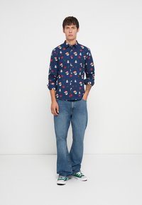 Camisa de botones navy con un estampado festivo de caras de Santa, copos de nieve y árboles; combinada con jeans de mezclilla claros y zapatillas verdes.