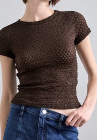 Braun strukturierte Bluse mit kurzen Ärmeln, die ein figurschmeichelndes Design und ein dezentes Muster aufweist. Kombiniert mit hoch taillierten blauen Jeans.