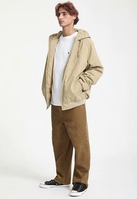 Beige lightweight jacket with a hood, paired with a white T-shirt and brown corduroy pants. Black low-top sneakers complete the outfit.