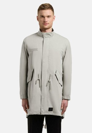 Mann iført en lys grå lang parka med høy krage, snorer i midje og nederkant, klafflommer og svarte bukser mot en ensfarget bakgrunn.