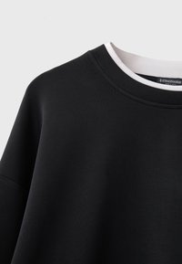 T-shirt noir à col rond superposé à un détail de col blanc, montrant la partie supérieure de la poitrine et l'épaule sur un fond uni clair.