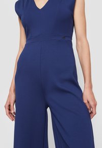 Femme portant une combinaison sans manches bleu marine avec un col en V et un pantalon à jambes larges sur un fond uni.