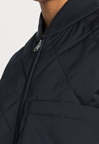 Giacca bomber trapuntata di colore navy con colletto a coste, chiusura con zip argentata e motivo di cuciture diagonali. Texture morbida e liscia.
