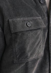 Chemise en velours côtelé gris foncé avec une poche poitrine à bouton, tissu texturé à motif côtelé vertical et coutures contrastantes.
