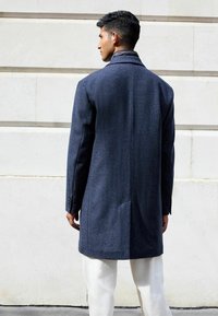 Marineblauer Wollmantel mit taillierter Passform, Reverskragen, strukturiertem Stoff und Seitentaschen. Die Rückansicht zeigt eine schlichte Silhouette.
