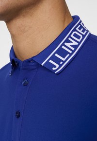 Blue polo shirt with a contrasting ribbed collar featuring white lettering. The fabric is textured with a smooth finish and button details.