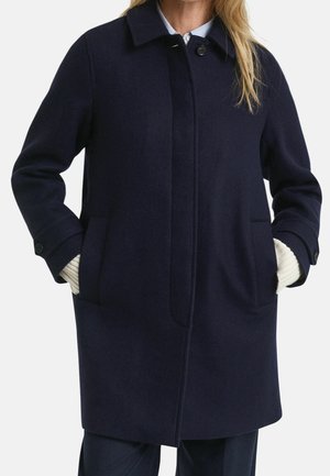 Woman wearing a navy blue wool coat with front pockets, buttoned cuffs, and a concealed button front closure over a light sweater.