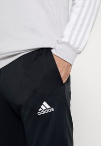 Maglietta grigia a maniche lunghe con tre strisce bianche sulla manica, abbinata a pantaloni sportivi neri con logo Adidas bianco.