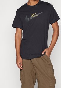 Černé bavlněné tričko s výrazným černým a zlatým logem Nike na hrudi, v kombinaci s khaki cargo šortkami se bočními kapsami.