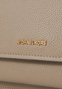 Bež kožna torba s teksturiranom površinom, koja sadrži zlatni "MICHAEL KORS" logo i kontrastnu šavovu uz rubove.