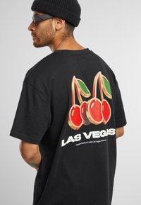 Sort bomulds T-shirt med et grafisk motiv af røde kirsebær med grønne blade og teksten "LAS VEGAS" i hvid, placeret på ryggen.