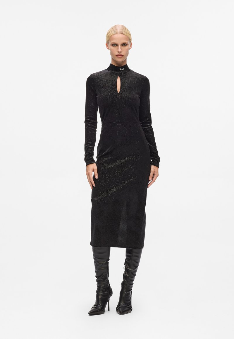 Vestido midi preto de mangas longas em tecido suave e texturizado, com detalhe de decote em forma de chave. Modelo usa botas pretas de salto alto e bico fino.