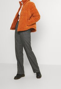 Veste matelassée en velours côtelé orange avec un col haut, associée à un pantalon en plaid gris et des chaussures noires à lacets. Textures plates et motifs distincts.