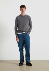 Mężczyzna stojący w pobliżu białej ściany, ubrany w szary sweter, niebieskie dżinsy i czarne buty, z jedną ręką w kieszeni.