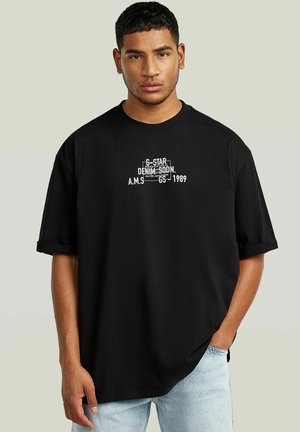Jeune homme aux cheveux courts et bouclés portant un t-shirt noir oversize avec un texte blanc et un jean bleu clair, une main dans la poche sur un fond uni.