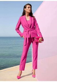Fuchsia-Anzug mit einem taillierten Blazer und Slim-Fit-Hosen, mit einem Gürtel in der Taille. Kombiniert mit passenden Absätzen und einer kleinen Handtasche.