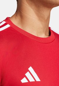 Rotes Sportshirt mit rundem Halsausschnitt, glatter Textur und weißem Adidas-Logo sowie drei weißen Streifen auf den Schultern.