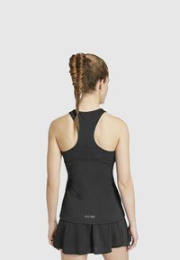 Vrouw met vlechten in het haar, gekleed in een zwarte racerback tanktop en een tennisrok, gezien vanaf de achterkant op een effen achtergrond.