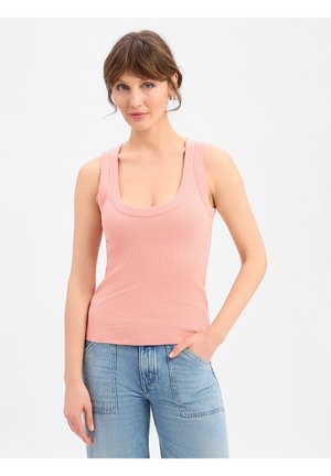 Vrouw met kort bruin haar, gekleed in een roze mouwloos shirt en lichtblauwe spijkerbroek, staand met één hand in haar zak tegen een effen achtergrond.