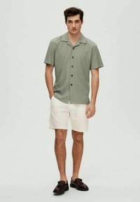 Camisa de botão de manga curta, verde-claro, com colarinho, apresentando uma textura suave. Combinada com calções bege e sapatos castanhos.
