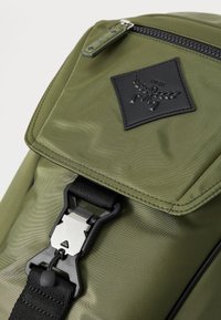 Rucsac din nailon verde măsliniu, cu un capac mare, accente din piele neagră și o cataramă din plastic argintiu. Dispune de o suprafață texturată și fermoare negre.